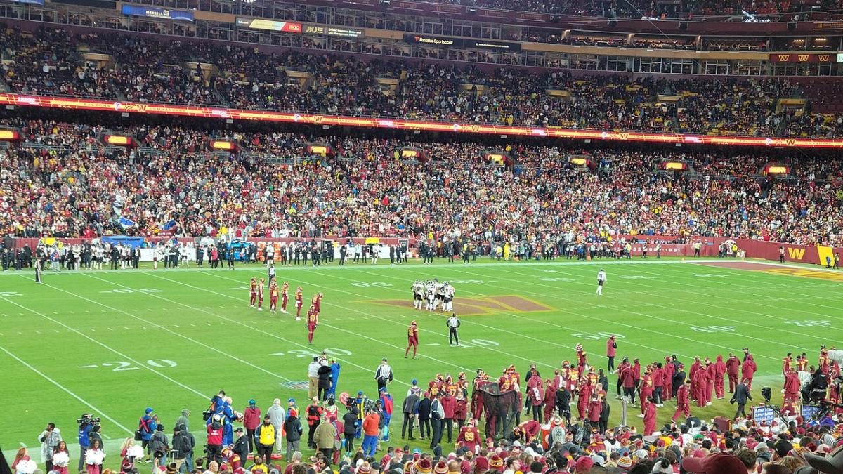 Washington players on the field at Northwest Stadium during a 2024 game, with the stands behind them.